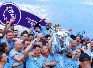 Manchester City manager Pep Guardiola and his players celebrate winning the Premier League title by lifting the trophy. The image shows the team in their sky blue kits surrounded by blue confetti, with a Premier League 'Champions 2022/23' graphic in the corner.
