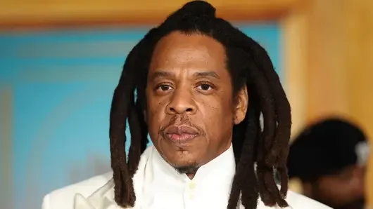 A medium close-up photograph of American rapper and businessman Jay-Z. He is shown with his signature long dreadlocks, wearing a crisp white dress shirt and matching white suit jacket against a blurred background.