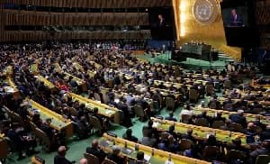 A high-angle photograph showing the United Nations General Assembly hall in New York filled with international delegates. A speaker stands at the green marble podium at the front, flanked by large screens displaying his image beneath the golden UN logo.