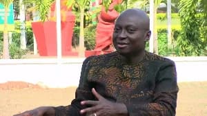 A portrait of Samuel Atta Akyea, a prominent Ghanaian lawyer and politician, seated outdoors. He is wearing a dark, patterned traditional shirt and appears to be speaking or gesturing during an interview or public appearance.