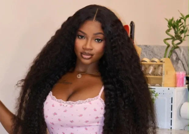 A portrait of a young woman identified as Adwoa Tee, featuring her with long curly hair and wearing a pink floral top. She is positioned in an indoor setting that resembles a kitchen, with household items like a spice rack and a bamboo plant visible in the background.