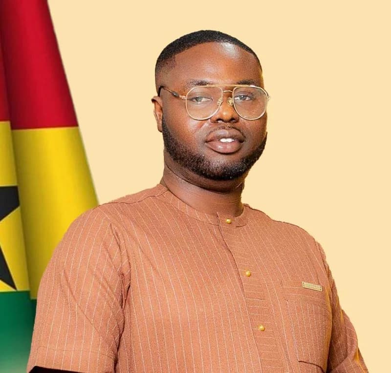 A formal portrait of Gabriel Tanko Kwamigah-Atokple, the newly appointed Patron of the NDC UK and Ireland Aldershot Branch. He is pictured wearing a brown pinstriped traditional Ghanaian outfit and glasses, positioned in front of the Ghana national flag.