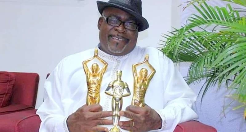 Ghanaian veteran actor Kofi Adjorlolo is pictured smiling while holding three gold award statuettes. He is wearing a white traditional outfit, a black hat, and spectacles, seated in an indoor setting with a red sofa and greenery in the background.