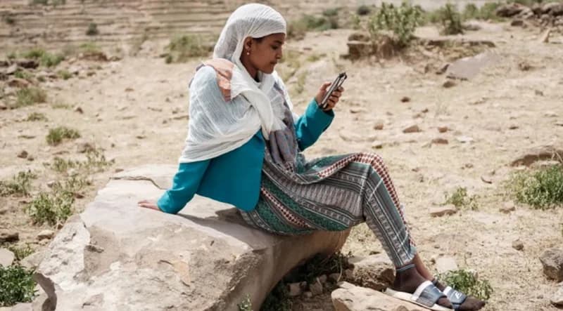 A woman wearing a white headscarf and a teal jacket sits on a large rock in a dry, rocky landscape while looking at her smartphone. The background features arid, terraced terrain typical of rural regions in Ethiopia.