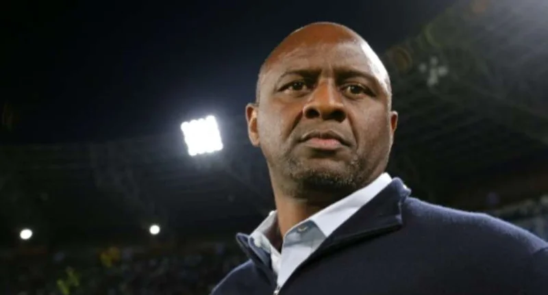 A close-up portrait of football coach Patrick Vieira at a stadium at night. He is wearing a dark blue zip-up sweater over a light blue collared shirt, looking forward with a serious expression against a background of stadium floodlights.
