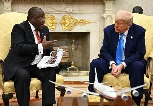 South African President Cyril Ramaphosa and Donald Trump are seated in yellow armchairs during a meeting in the Oval Office. Ramaphosa is holding documents and speaking while gesturing towards Trump, who is listening attentively.