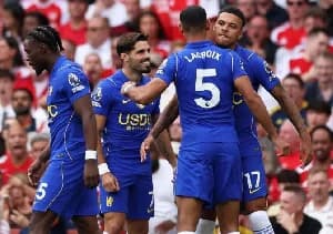 Chelsea football players, including Maxence Lacroix and Jadon Sancho, are seen celebrating on the field during a match. The players are wearing their blue home kits with the USDC sponsorship visible, set against a background of fans in the stadium stands.