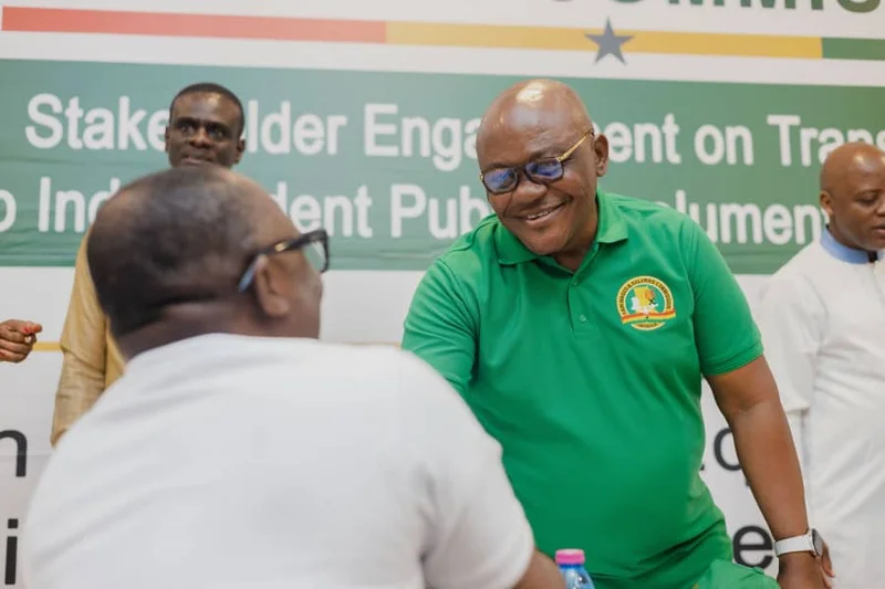 A man wearing a green Fair Wages and Salaries Commission (FWSC) polo shirt smiles while interacting with an attendee at a stakeholder meeting. A banner in the background mentions 'Stakeholder Engagement on Transition to Independent Public Emoluments', indicating a government or policy discussion.