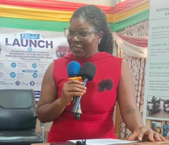 A woman in a red sleeveless dress and glasses is captured giving a speech at the official launch of the WASH and Livelihood Improvement Project. She is holding two microphones at a podium, with a project banner and Ghana-flag-colored drapes visible in the background.