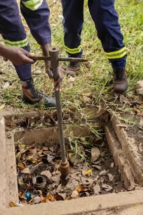 Two fire service personnel in blue uniforms with reflective yellow stripes are pictured inspecting an underground fire hydrant. One officer is using a long metal T-handle wrench to operate a valve located inside a concrete pit filled with dry leaves and debris.