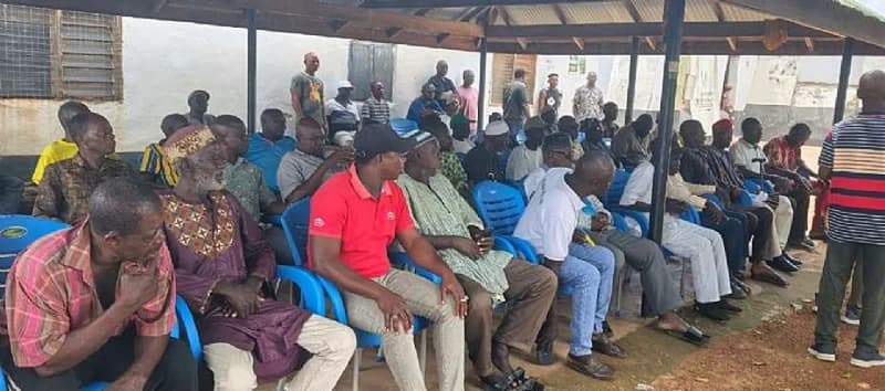 A group of men are gathered under a pavilion for a community meeting, seated on blue plastic chairs. The attendees, including several elders in traditional Smocks, appear to be engaged in a discussion regarding local infrastructure and road construction in Tumu.