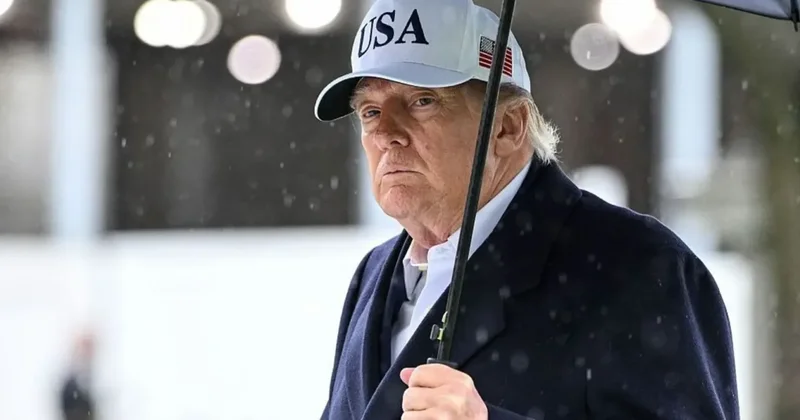 A close-up photograph of Donald Trump wearing a white cap with 'USA' text and an American flag emblem. He is dressed in a dark navy overcoat and is holding a black umbrella while outdoors in what appears to be rainy or snowy weather conditions.