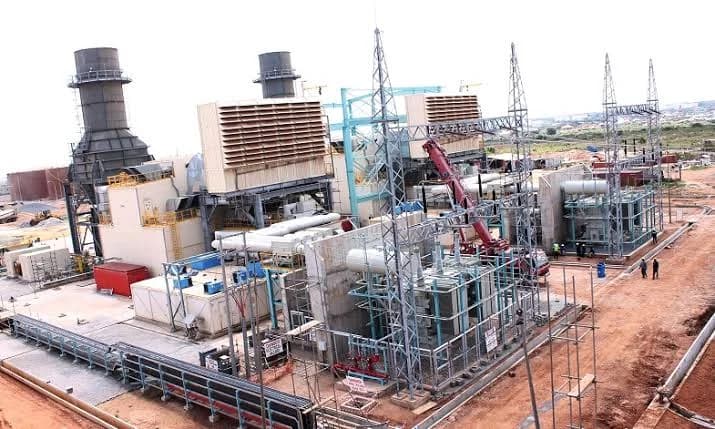 A wide-angle photograph of a large gas power plant facility, featuring multiple exhaust stacks and extensive electrical infrastructure. The site appears to be an active industrial zone with various technical structures, cooling units, and transmission towers under a bright sky.