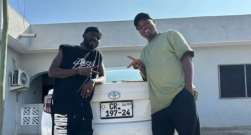A photograph showing Ghanaian artist Orkortor Perry and his producer 420 Drumz posing behind a white Toyota Camry. Both men are smiling and making peace signs, standing in front of a white building with a visible Ghanaian license plate.
