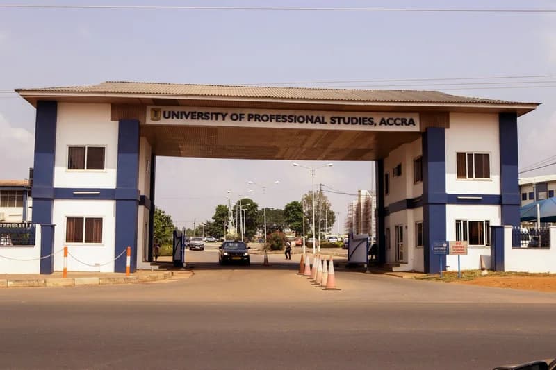 The image displays the prominent main entrance gate of the University of Professional Studies, Accra (UPSA). It features a large blue and white archway with the university's name clearly displayed across the top, overlooking an asphalt road with light traffic and security bollards.