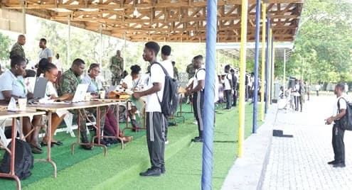 Young recruits stand in line at an outdoor documentation center while officials and military personnel process their information using laptops. The scene captures a selection exercise for the Ghana Armed Forces, featuring individuals in civilian attire and military camouflage under a large wooden structure.
