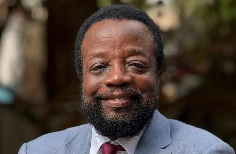 A headshot portrait of Kofi Koranteng, the flagbearer of the Progressive Alliance of Ghana (PAG). He is seen smiling, wearing a grey suit jacket, a white shirt, and a maroon patterned tie against a blurred outdoor background.