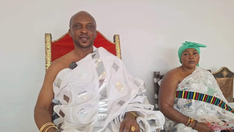 A traditional Ghanaian leader, likely the Chief of Yefri, sits on a red-backed throne wearing a white patterned cloth and gold jewelry. Beside him sits a woman in traditional white lace and a green headwrap. The man has a lapel microphone attached to his garment, suggesting he is giving a formal address or interview regarding the community's harvest and needs.