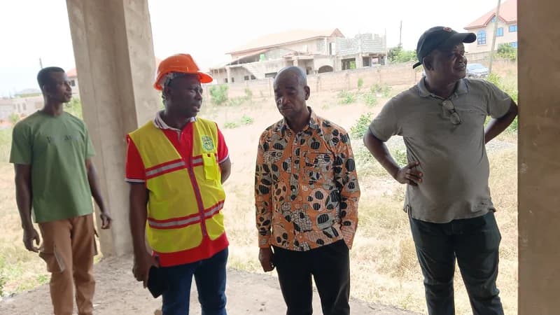 A group of men, including a project official in a high-visibility safety vest and hard hat, inspect the construction progress of a building in Kpone-Katamanso. They are standing within the concrete shell of the unfinished structure, which is part of the new 24-hour market project.