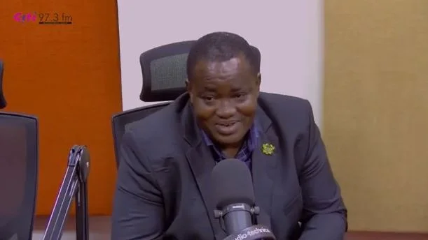 A photograph of Dr. Henry Kwabena Kokofu, a prominent Ghanaian official, speaking during a media interview at Citi FM. He is wearing a dark suit and is positioned in front of an Audio-Technica microphone within a radio studio setting.