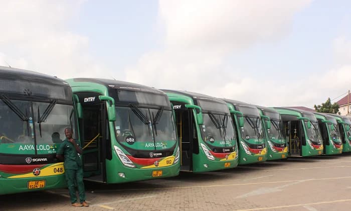 A row of green and black Aayalolo buses, branded with the Scania logo, are lined up in a parking area in Ghana. A man wearing a dark green uniform leans against the front bus on the left, while the fleet stretches into the background under a pale sky.