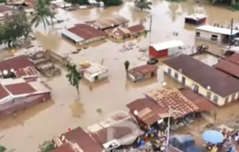 An aerial photograph showing a residential neighborhood in Hohoe severely impacted by flooding. Numerous buildings with red-tiled and corrugated metal roofs are partially submerged in murky, brown water that has covered streets and yards.