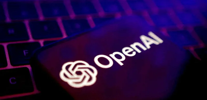 A close-up photograph featuring a smartphone displaying the OpenAI logo and wordmark, positioned atop a laptop keyboard. The image is characterized by a high-contrast, atmospheric lighting scheme using purple and blue tones, symbolizing modern digital technology.