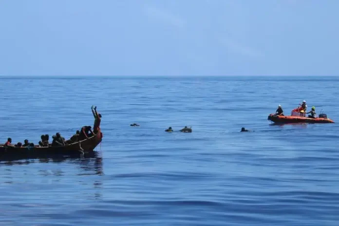 A wide shot shows a maritime rescue operation in the Mediterranean Sea involving an overcrowded wooden boat and an orange inflatable rescue vessel. Several individuals are visible in the water between the two boats, with one person on the wooden craft raising their arms in a gesture for help.