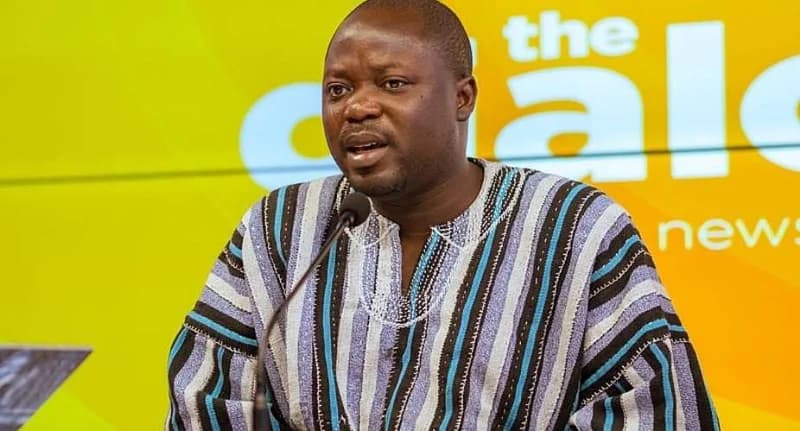 A man wearing a traditional Ghanaian smock is captured speaking into a microphone during what appears to be a television interview or press briefing. Behind him is a vibrant yellow and green background with the words 'the news' partially visible, indicating a media setting.