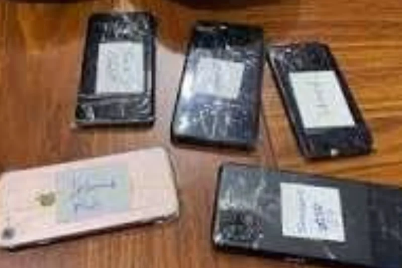 The image shows five smartphones laid out on a wooden surface, several with visible cracks on their screens. Each device has a small white label with handwritten notes attached, suggesting they have been collected as evidence in a legal or criminal investigation.