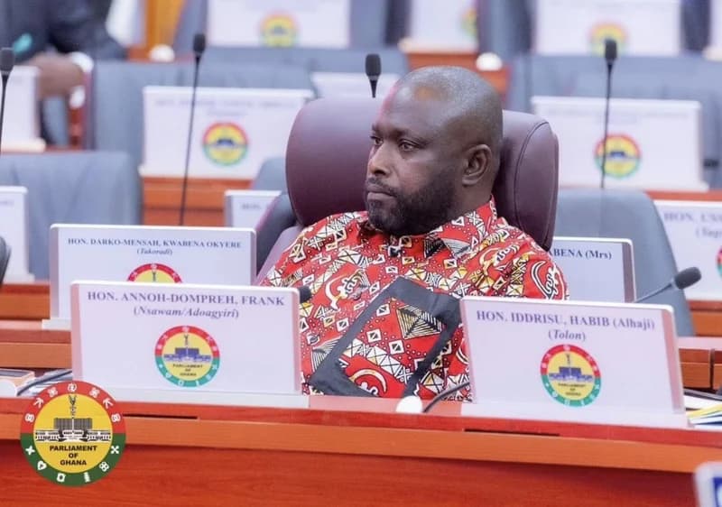 Hon. Habib Iddrisu, Member of Parliament for Tolon, is seen seated in the Parliament of Ghana chamber. He is wearing a red patterned shirt and sitting behind his official nameplate, surrounded by the nameplates of other Members of Parliament.