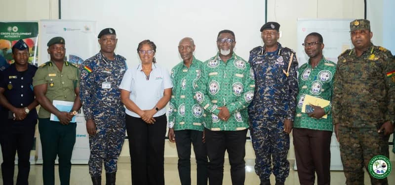 A group of nine individuals, including security personnel and officials, pose for a group photograph indoors. The group features members of the Ghana Police Service and military in uniform alongside civilians wearing branded Tree Crops Development Authority (TCDA) shirts.