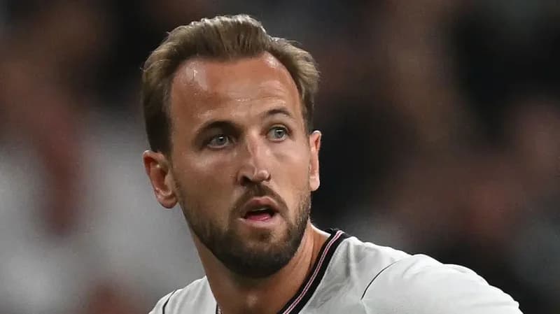 A close-up photograph of English footballer Harry Kane looking off-camera with a focused expression. He is wearing a white England national team training or match jersey with a thin red and black collar.