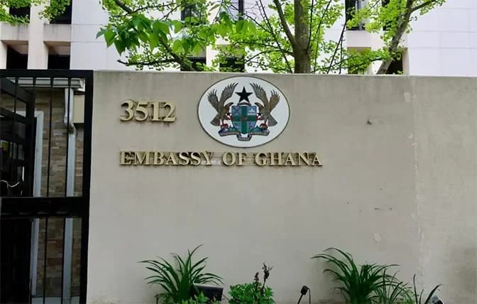 The image shows the exterior entrance of the Embassy of Ghana, featuring the official Coat of Arms and the words "EMBASSY OF GHANA" in gold lettering on a tan wall. Green foliage and parts of the embassy building are visible in the background.