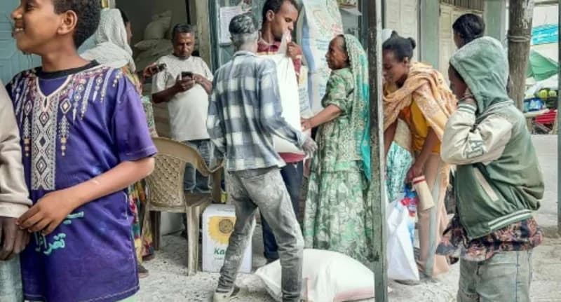 A group of people are gathered at a distribution point or marketplace in a region likely affected by the Ethiopia-Eritrea conflict. Men are seen carrying large white sacks of food supplies, while others, including women in traditional dress and a child in a patterned purple shirt, observe the distribution process.