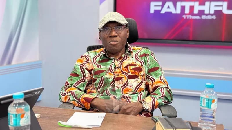 Former Member of Parliament Inusah Fuseini is seated in a television studio, looking directly at the camera. He is wearing a colorful patterned shirt, glasses, and a flat cap, with a notepad and water bottles positioned on the desk in front of him.
