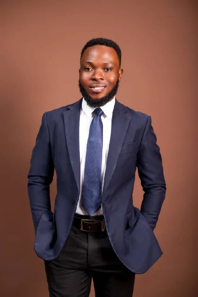 A formal studio portrait of a young man, likely Samuel Kofi Payitey, posing against a solid brown background. He is dressed in professional attire consisting of a navy blue blazer, a white shirt, and a blue necktie, looking directly at the camera with a confident smile.