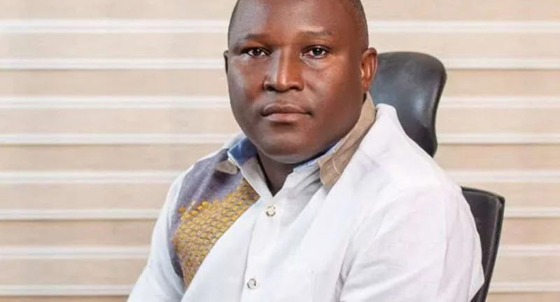 A medium-shot portrait of Daryl Bosu, Deputy National Director of A Rocha Ghana, sitting in a black office chair. He is wearing a white collared shirt with a grey and gold patterned detail on the right side, set against a background of neutral-toned horizontal blinds.