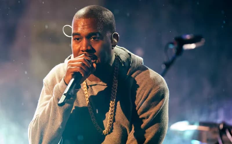 American rapper Kanye West is pictured performing on stage, holding a microphone and wearing a grey hoodie with a thick gold chain. The photograph captures him in a moment of performance with atmospheric stage lighting and haze in the background.
