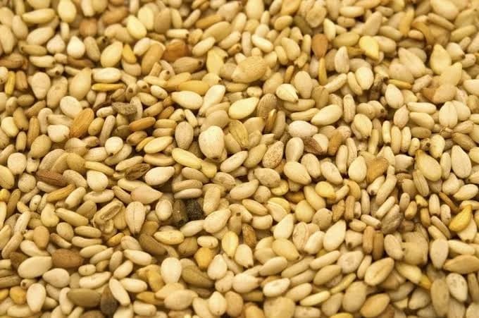 A high-detail close-up photograph showing a dense collection of sesame seeds. The seeds display natural variations in color ranging from off-white and cream to light brown.