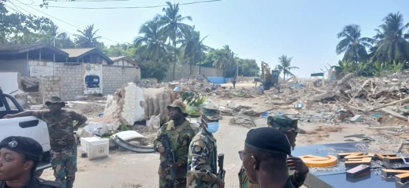 The image shows Ghana Armed Forces personnel in camouflage uniforms standing guard at a demolition site, likely at La Beach in Accra. In the background, an excavator is visible amidst the rubble of destroyed buildings and piles of debris, with palm trees lining the horizon. The scene depicts the aftermath of a demolition operation with a significant military presence.