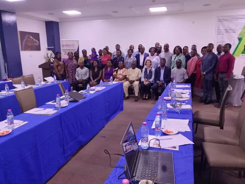 A group photograph of professionals attending a technical working group meeting in a conference room. Participants are arranged behind U-shaped tables covered with blue cloths, featuring Verna water bottles and laptops, with INSLA organizational banners visible in the background.