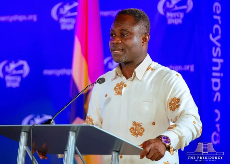 Kwabena Mintah Akandoh, Ranking Member on the Parliamentary Select Committee on Health, is seen speaking at a podium. The backdrop features Mercy Ships branding and a Ghanaian flag, indicating an official event related to the organization's mission in Ghana.
