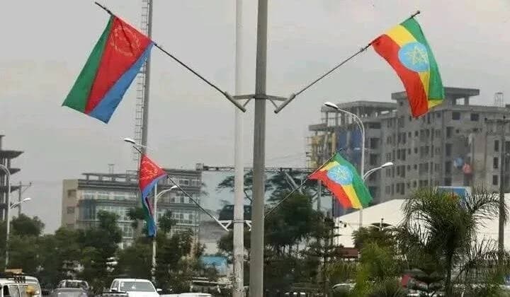 A photograph showing the national flags of Eritrea and Ethiopia flying side-by-side on street poles in an urban area. The scene features multi-story buildings and vehicles in the background under an overcast sky, representing the diplomatic connection between the two nations.