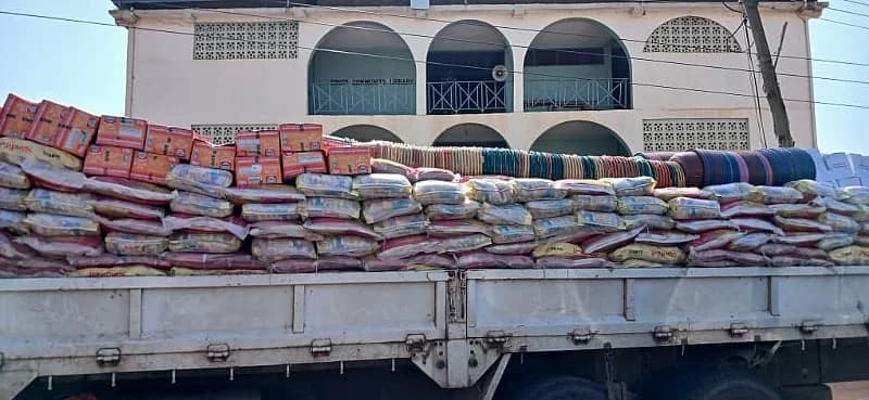 A large truck is heavily loaded with relief items including stacked sacks of food, boxes, and colorful plastic basins. The vehicle is parked in front of the Hohoe Community Library building, which features distinctive arched balconies.
