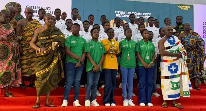 Students from Weweso M/A JHS pose with a trophy and medals alongside traditional leaders at the Otumfuo Community Reading Challenge. The winners, dressed in green and yellow branded polo shirts, stand on a red-carpeted stage in front of an event backdrop. Several chiefs in ceremonial Kente cloth are present to support the educational initiative.