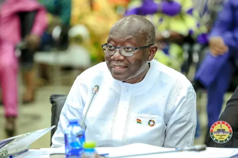 A photograph of Ken Ofori-Atta, Ghana's former Minister for Finance, seated at a formal gathering, likely within the Parliament House. He is wearing a white kaftan with visible Ghana flag and Sustainable Development Goals (SDG) pins, looking toward someone off-camera with a slight smile.