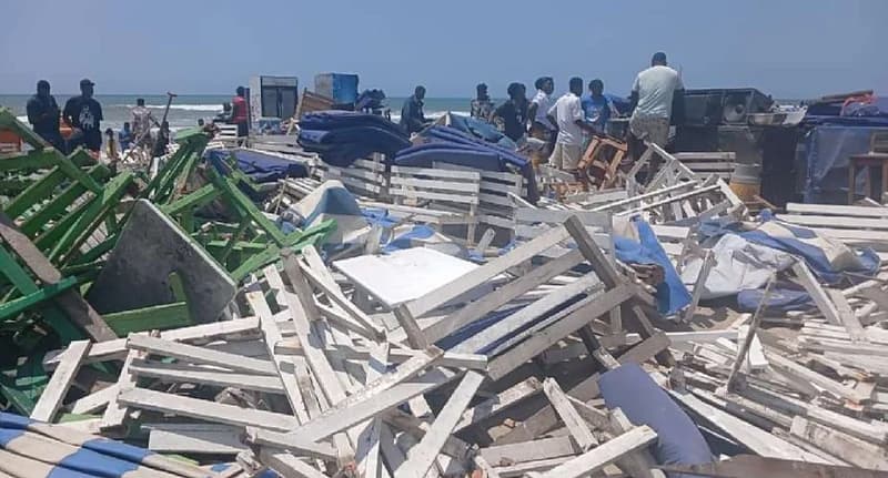 The photograph captures the aftermath of a demolition exercise at Labadi Pleasure Beach, showing large piles of broken white and green wooden benches and tables. A group of people stands in the background amidst more debris, with the ocean and clear blue sky visible behind them.