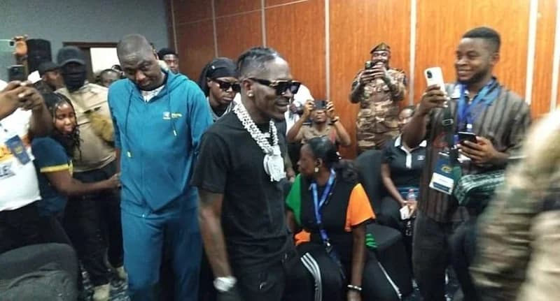 Ghanaian dancehall artist Shatta Wale, wearing a large diamond-encrusted chain and dark sunglasses, is captured at an indoor event surrounded by fans and press. Several individuals in the background are holding up smartphones to record or photograph the artist, who is dressed in a black t-shirt.