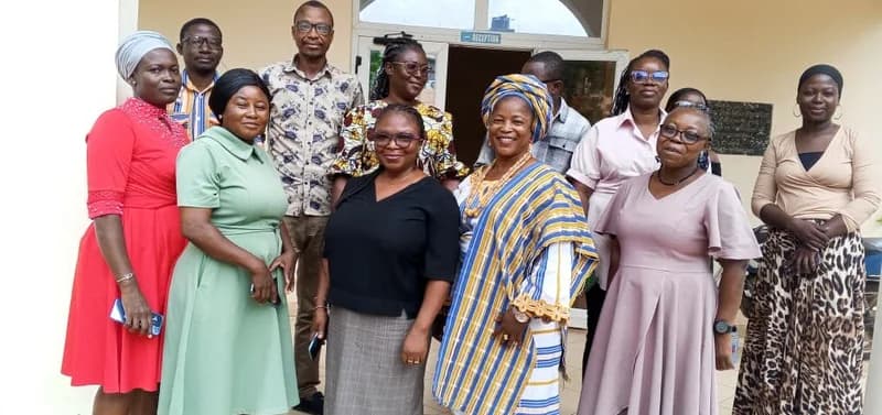 A group of stakeholders, primarily women, pose for a group photograph in front of a building entrance labeled 'RECEPTION'. The individuals are dressed in a mix of Western and traditional Ghanaian attire, with one woman prominently featured in a blue and white kente-style wrap.